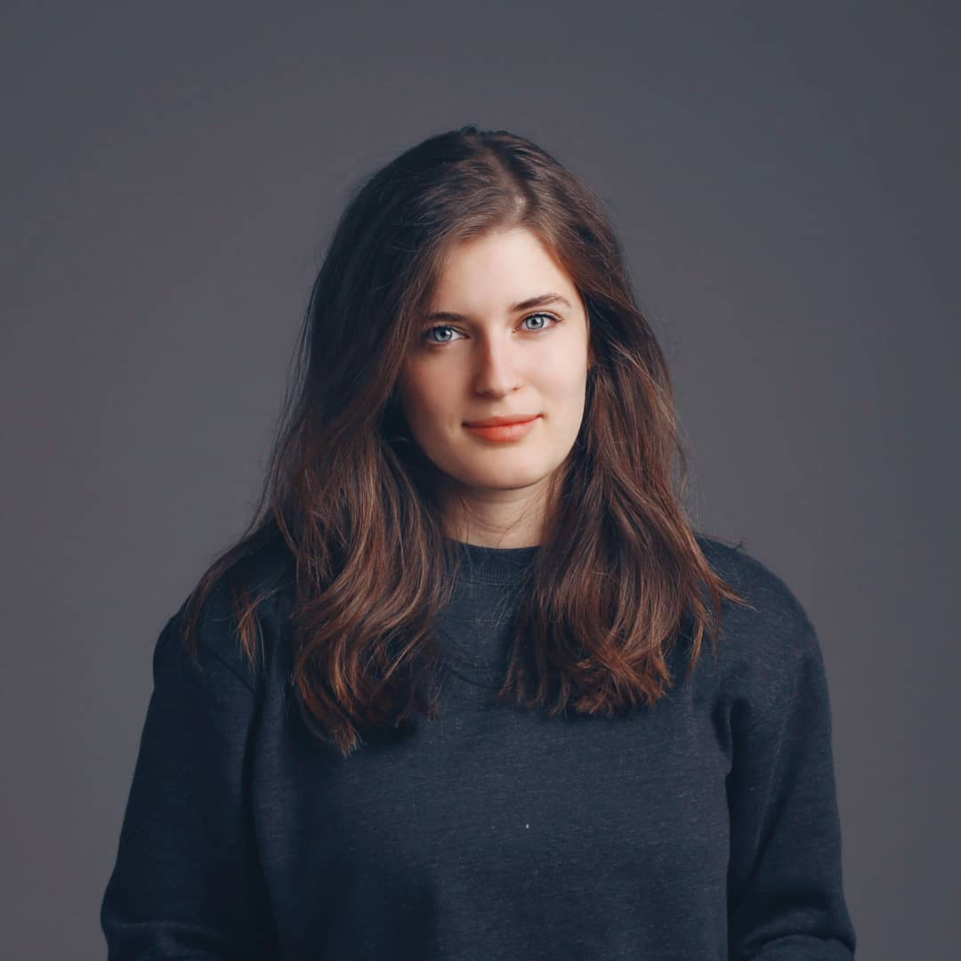 Shkendie: White girl with blue eyes and brown hair wearing a dark blue sweater in front of a dark background