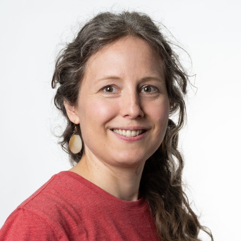 Person with long wavy brown hair wearing a red crew-neck shirt and mother-of-pearl earrings, photographed against a plain white background.